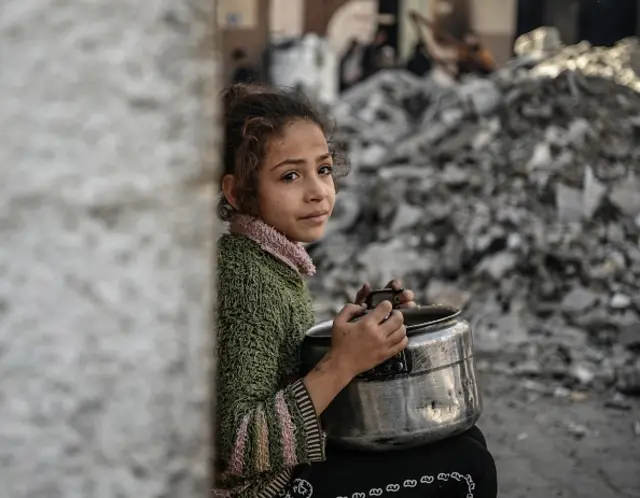 A Palestinian child, holding empty pot, waits near rubbles to receive food distributed by volunteers, 22 December 2023