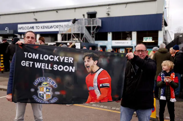 Fans pose for a photo whilst holding a banner displaying the message "Tom Lockyer get well soon" prior to the Premier League match between Luton Town and Newcastle United