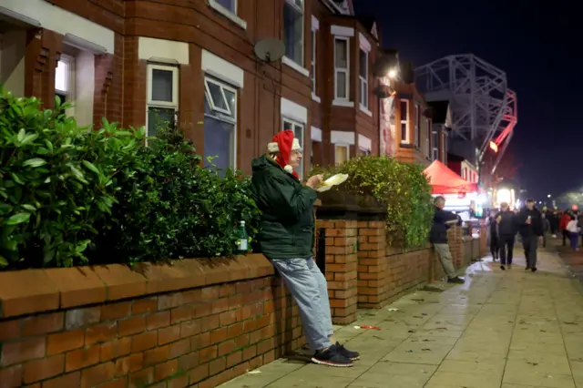 A fan with a Santa hat sits on a front wall eating chips with Old Trafford in the background.