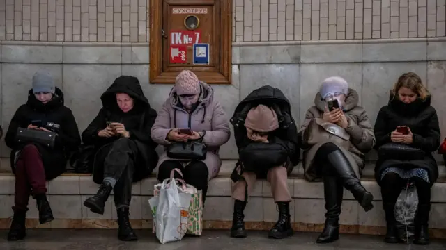 Women sitting looking at their phones. One has her head down, in her hands, as if sleeping or crying.