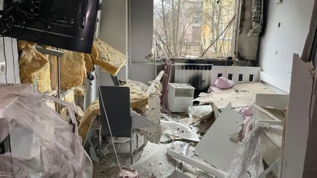 Parts of a wall scattered over bedroom with windows blown out and black smoke damage around the windo