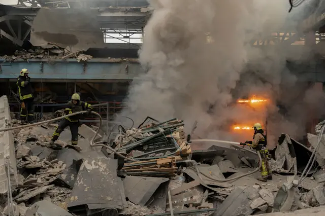 Firefighters spray water on fire, standing on huge piles of debris in front of destroyed buliding