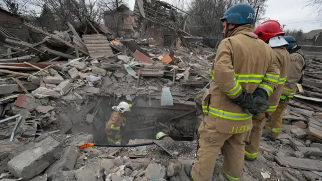 Ukrainian emergency workers stand in and amongst the ruins of buildings