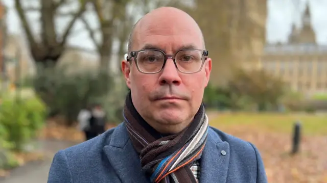 Steve Brisley in a blue blazer and a scarf with a solemm expression