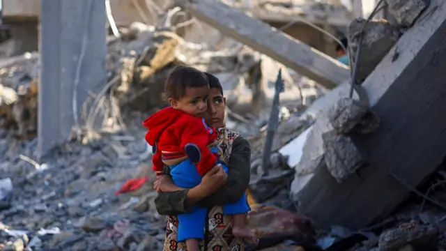 A boy holds a baby, looking up at the destruction around him