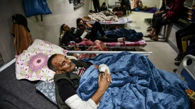 in focus, a man sleeps in a hospital bed in a white-tiled hallways. Behind him are many others