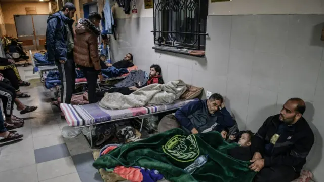 Camp beds line a hallway. In the foreground, two men comfort two sleeping children