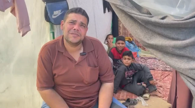 Mohamed sits on the floor, looking up at the camera and wearing a red polo shirt. Small children sit behind him on the floor under what appears to be a makeshift tent.