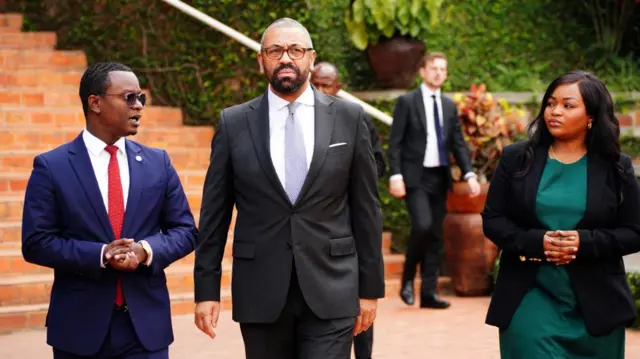 Cleverly walks along an orange tiled path, flanked by two others in suits