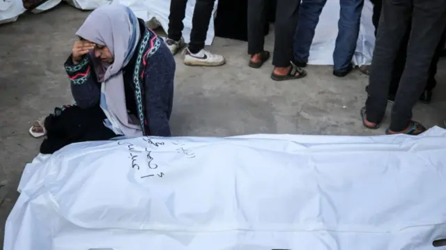 A woman holds her head despairingly as she sits on the ground next to a body bag