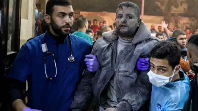 Man being held by two doctors. He is covered in grey ash and has blood on his face and jacket.