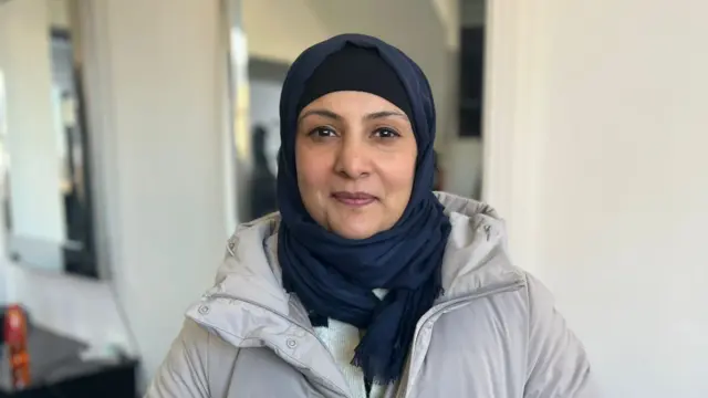 Sheetal. Sheetal wears a navy blue hijab and a grey puffer coat. She has brown eyes lined with black and smiles while looking at the camera. She is pictured inside against a white wall with a mirror behind her