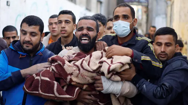 Man carrying child casualty in blankets, surrounded by other men