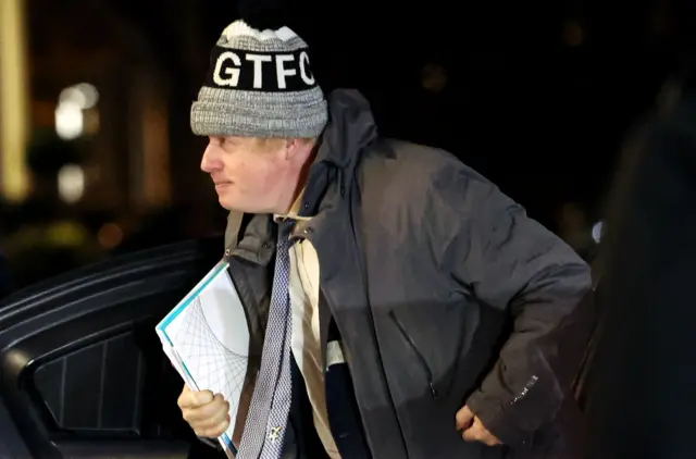 Boris Johnson getting out of a car holding a folder, wearing coat and beanie