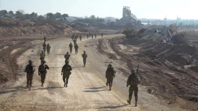 IDF soldiers seen on foot in Gaza. Around two dozen soldiers are seen walking along a stretch of road. Rubble can be seen on either side of the road.