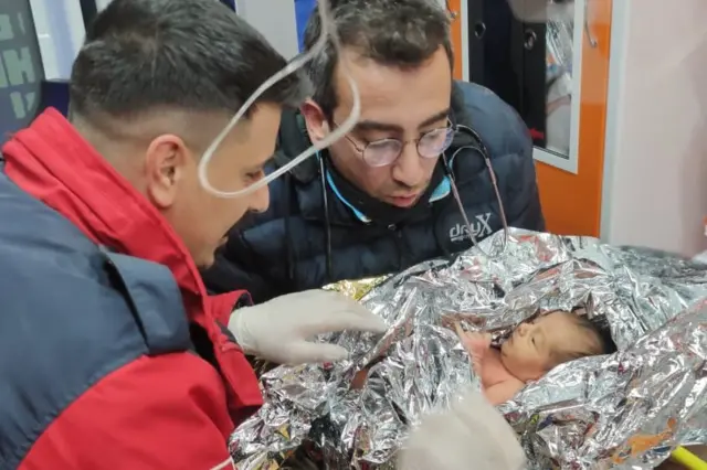 A baby wrapped in a thermal blanket is attended by two men after being rescued from a ruined building