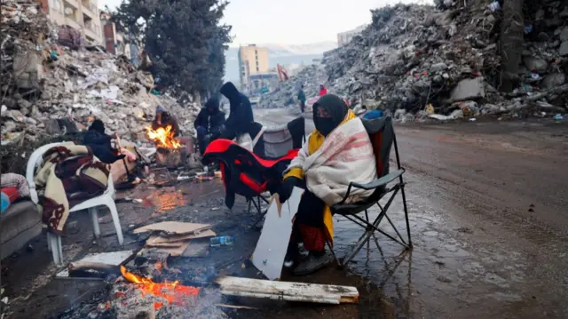 Relatives warm up by a fire among collapsed buildings in Kahramanmaras