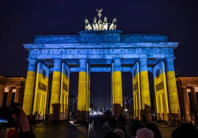 People stand in front of the Brandenburg Gate as it is illuminated in the colors of the Ukrainian flag