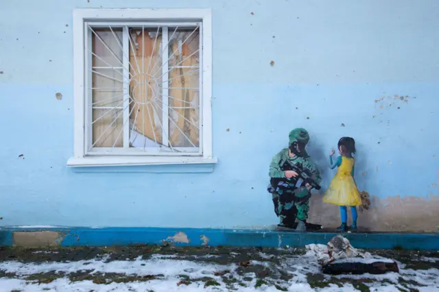 An artwork depicts an armed soldier kneeling next to a girl wearing blue and yellow