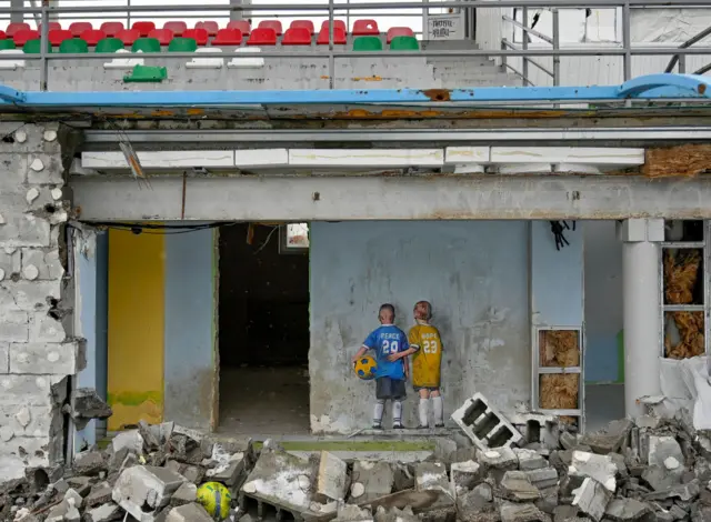 A mural of two boys, wearing blue and yellow, with a football, on the wall of a damaged stadium