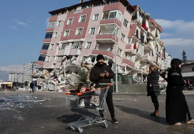 Children sit in a shopping cart near a collapsed building following an earthquake in Hatay, Turkey, February 7, 2023. REUTERS/Umit Bektas