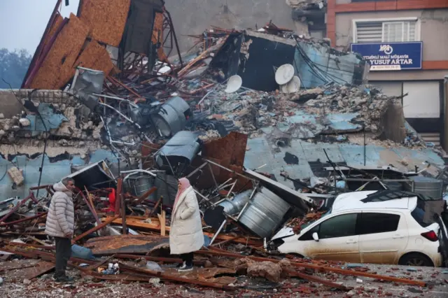 A man and a woman look at a collapsed building and a smashed car in Hatay, south-eastern Turkey. Photo: 7 February 2023