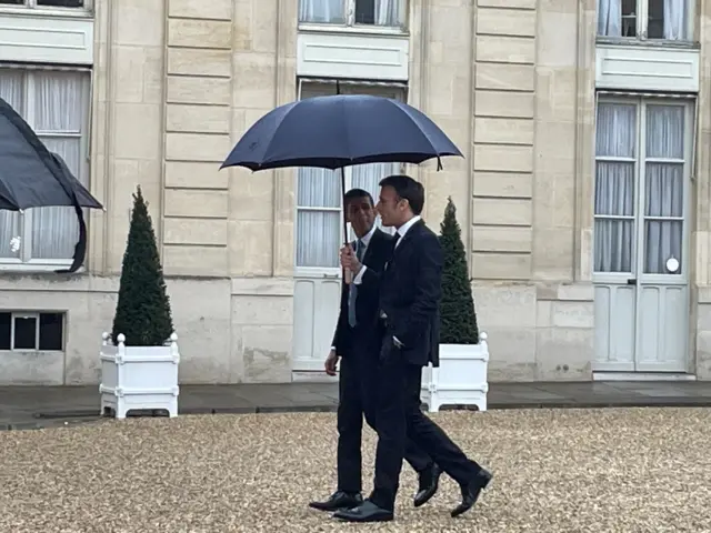 Emmanuel Macron carries an umbrella for Rishi Sunak as the two men walk together