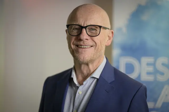 John Caudwell pictured in 2021. He is wearing glasses, a suit without a tie, and smiling at the camera.