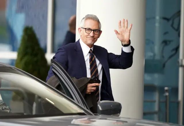 Gary Lineker wearing glasses and a suit, waving as he gets out of a car at Leicester's King Power stadium