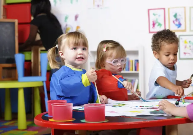 A portrait of a group of toddlers painting while their carer tidies.