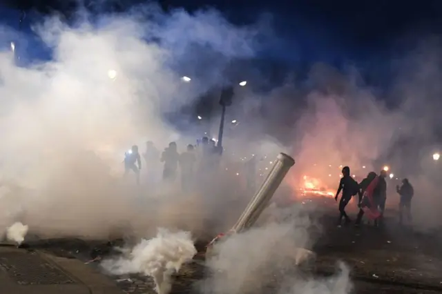 A group of unidentifiable people stand near a fire with tear gas around them