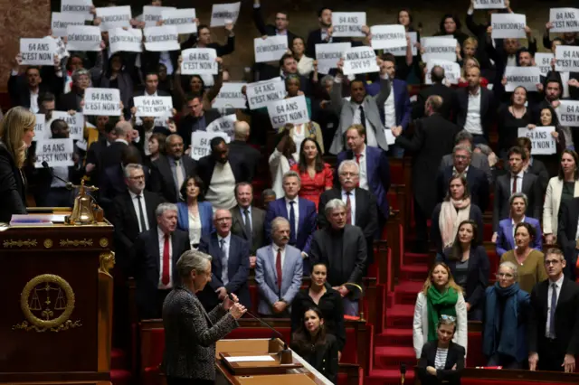 Elisabeth Borne speaking at a podium while opposition politicians hold up protest signs