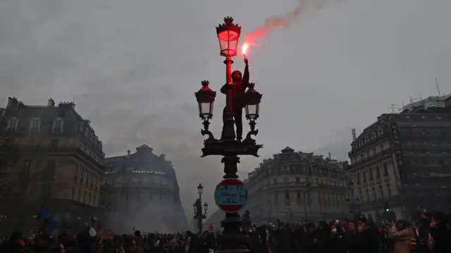 Protester standing on a lamppost lights a flare above a crowd of protesters