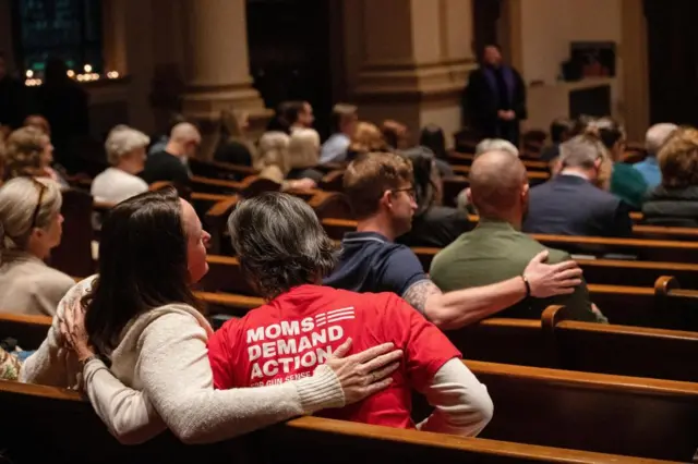 Mourners sit on pews at a vigil, one woman wears a tshirt which reads "Moms demand action for gun sense"