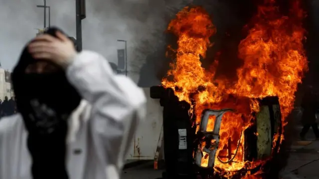 A masked protester stands in front of a burning car during clashes at a demonstration as part of the tenth day of nationwide strikes and protests against French government's pension reform in Nantes, France, March 28, 2023.