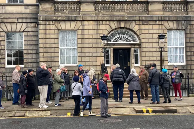 Tourists on a walking tour of Edinburgh are slightly bewildered by the gathering media outside the First Minister’s official residence.