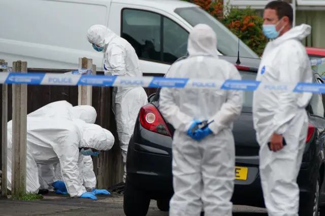 People in white suits and masks examine the scene behind police tape