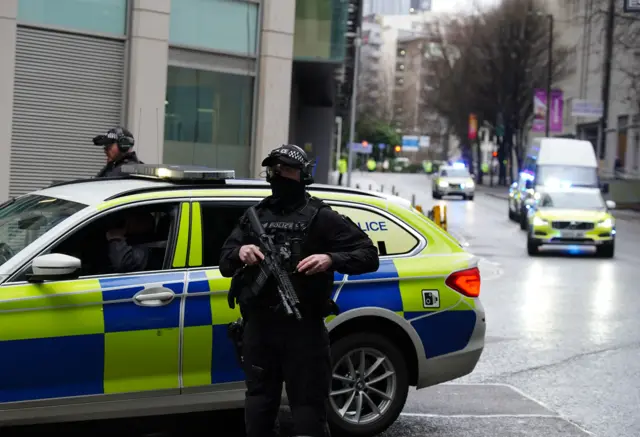 Two armed officers stand either side of a police car, as an escort approaches from behind