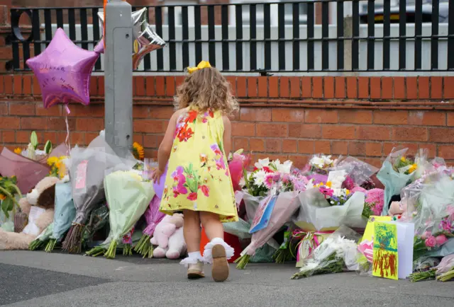 A girl in a yellow flowery dress leaves a tribute in front of a row of dozens of plastic-wrapped bouquets