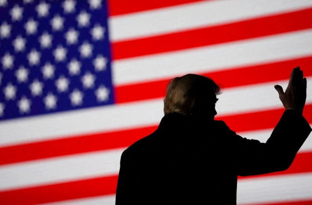 Donald Trump silhouetted on a stage against a screen showing the American stars and stripes flag