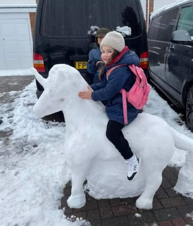 A young girl sits on a unicorn made out of snow in front if a vehicle on a driveway