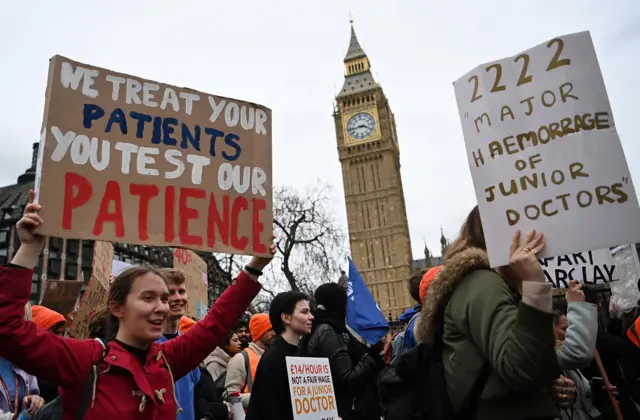 A woman holds a placard in a crowd outside Big Ben. The sign reads "we treat your patients, you test our patience"