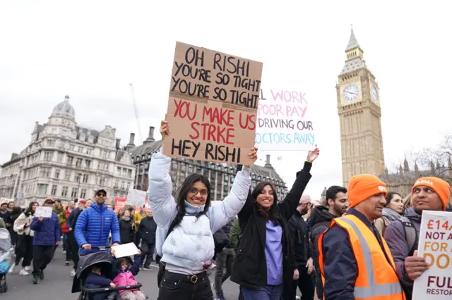 A woman holds a sign saying "Oh Rishi you're so tight, you're so tight you make us strike, hey Rishi" in front of Big Ben