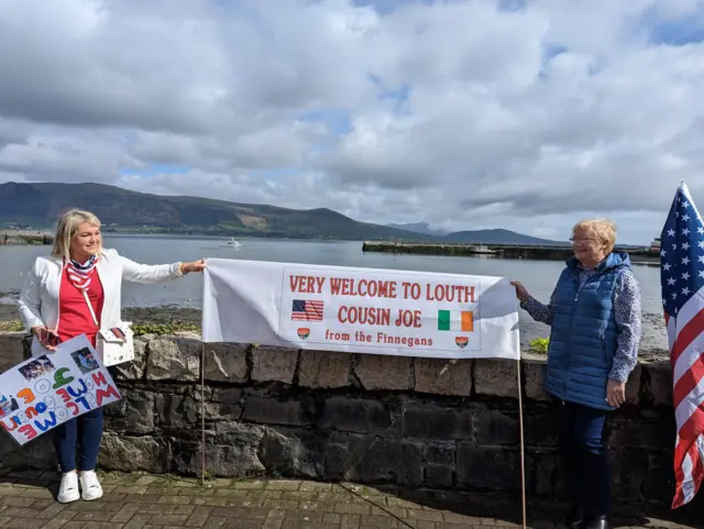 Two women hold a sign saying "very welcome to Louth, cousin Joe"