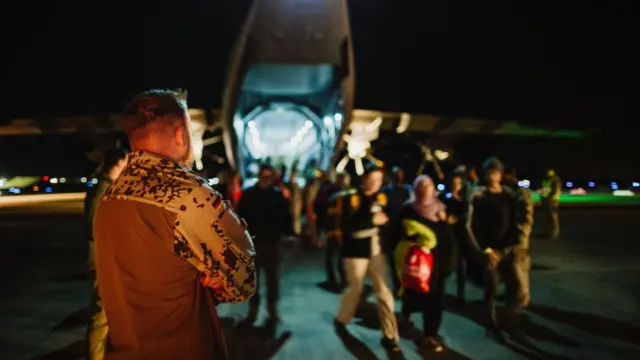 A military officer stands on a runway and watches people leave a plane