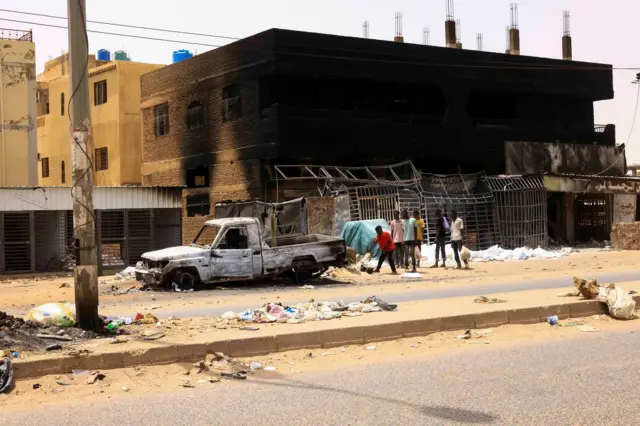 People walk near damaged car and buildings at the central market during clashes between the paramilitary Rapid Support Forces and the army in Khartoum North