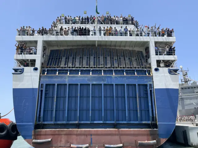 People wave from the deck of a ship as it reaches port