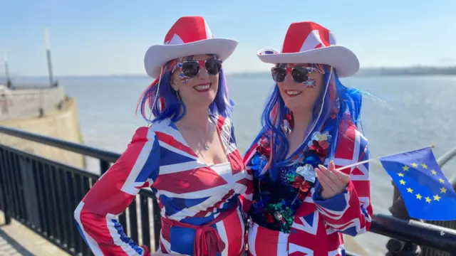 Two women wearing Union Jack dresses stand near the ocean