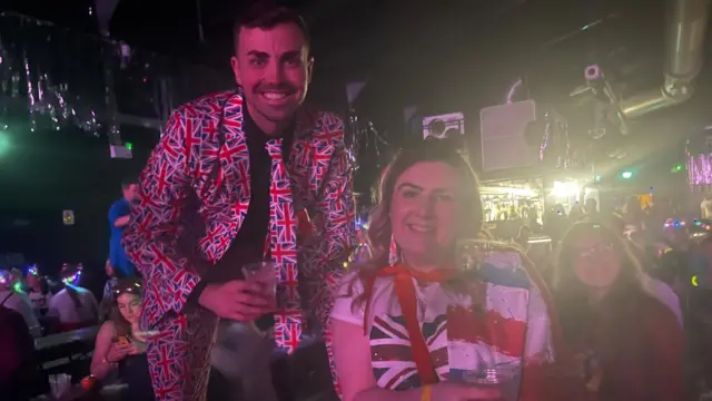 A woman stands with her brother on benches at a watching party