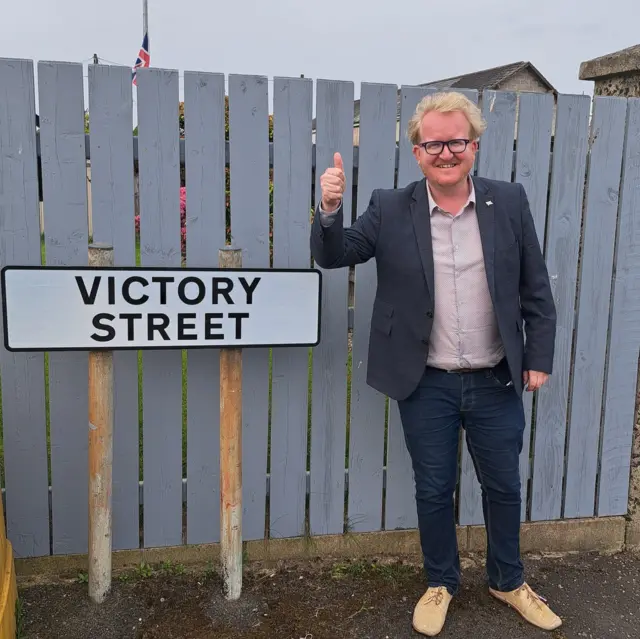 Gary Hynds poses besides a street sign that reads: Victory Street